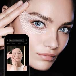 Junge Frau mit blauen Augen und innovativer Hautbehandlung, Smartphone zeigt digitale Schonheitsanwendung.