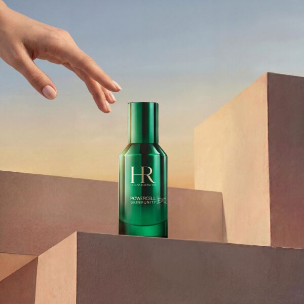 Produktfoto des Helena Rubinstein Powercell Skinmunity Exo Serums in einer glanzenden grunen Glasflasche. Die Flasche steht auf einem Stapel rosafarbener geometrischer Podeste, wahrend sich von oben eine elegante Hand nahert, vor einem warmen, sonnenuntergangsfarbenen Himmelshintergrund.​