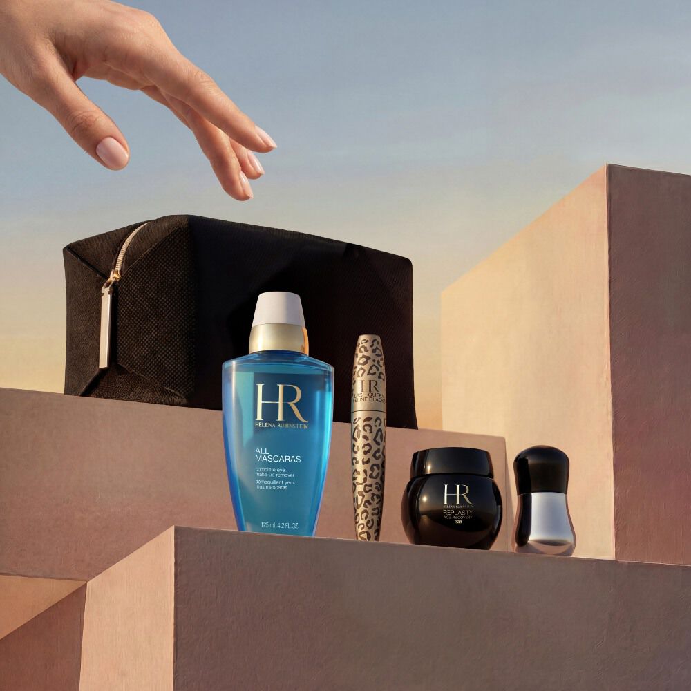 Eine elegante Hand greift nach einer sorgfaltig zusammengestellten Auswahl an Helena Rubinstein Produkten, die auf minimalistischen, sandbeigen Steinblocken angeordnet sind. Das Set wird vor einem warmen, goldenen Sonnenuntergangshimmel prasentiert und umfasst ein schwarzes Reise-Etui, den blauen Augen-Make-up-Entferner 'All Mascaras', eine Lash Queen Feline Blacks Mascara mit Leopardenmuster, einen kleinen schwarzen Re-Plasty Cremetiegel und ein silbernes Applikator-Tool.​