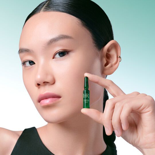 Nahaufnahme des Gesichts einer Frau, die eine grune Powercell [EXO] Skinmunity Ampulle von Helena Rubinstein delikat zwischen den Fingern halt, vor einem grun-weißen Farbverlaufshintergrund.​