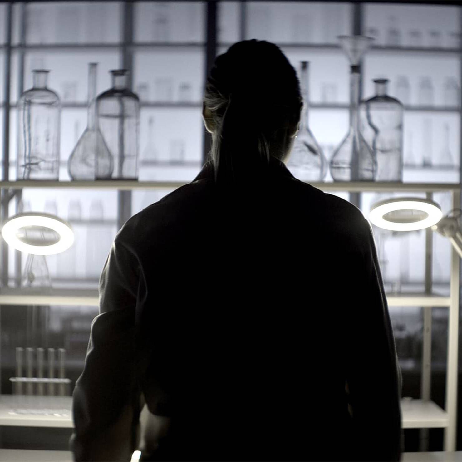 A silhouetted figure stands in a dimly lit lab, surrounded by various glass containers and illuminated by ring lights.