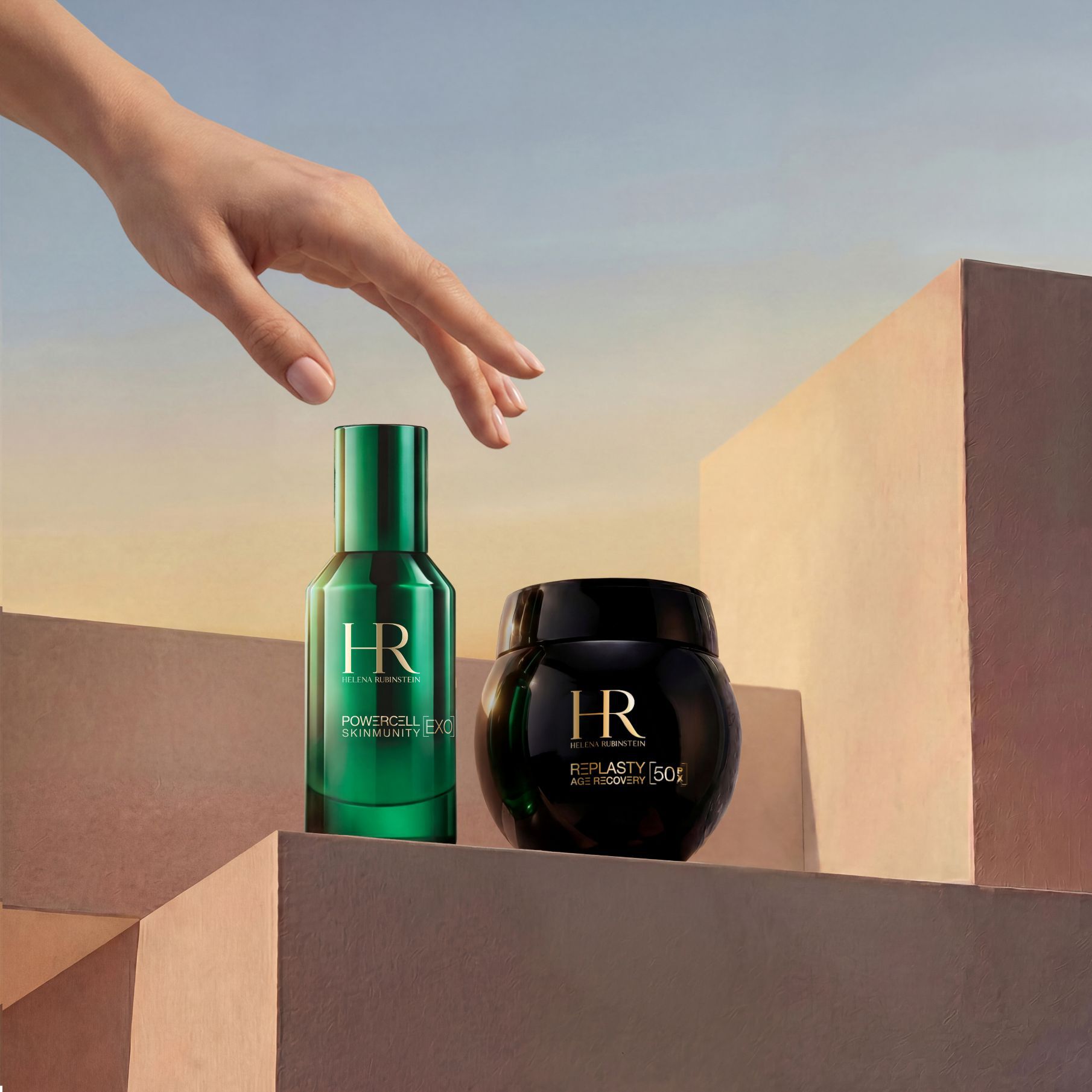 Eine elegante Hand greift nach dem Helena Rubinstein Duo Travel Replasty Night – Powercell Essence, bestehend aus einer grunen Flasche Powercell Skinmunity Essence und einem Tiegel Re Plasty Age Recovery Night Cream, prasentiert auf sandfarbenen geometrischen Blocken vor einem warmen Sonnenuntergangshimmel.​