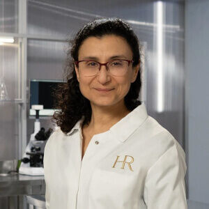 A researcher in a lab coat smiles confidently, with a microscope and lab equipment visible in the background.