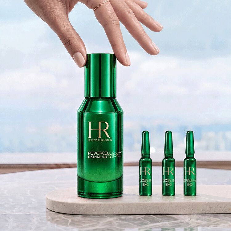 Eine Hand beruhrt die grune Flasche des Powercell [EXO] Skinmunity Serums von Helena Rubinstein, das neben drei [EXO] Shots Ampullen auf einem Marmorsockel vor einer hellen Landschaft steht.​