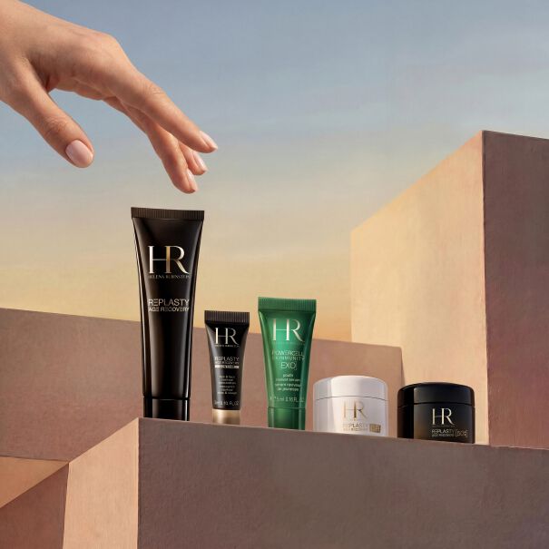 Produktfoto des monatlichen Geschenksortiments von Helena Rubinstein. Unter einem sanften Himmel mit Farbverlauf streckt sich eine zarte Hand nach Luxus-Pflegeprodukten aus, die auf minimalistischen, sandfarbenen Podesten prasentiert werden. Das exklusive Set umfasst einen Powercell Skinmunity Glasflakon, eine Powercell Skin Rehab Reisetube, zwei Powercell Skinmunity Exo Reisetuben, einen Tiegel Re-Plasty Age Recovery Day Cream [GR] und eine elegante weiße Kosmetiktasche im Hintergrund.​