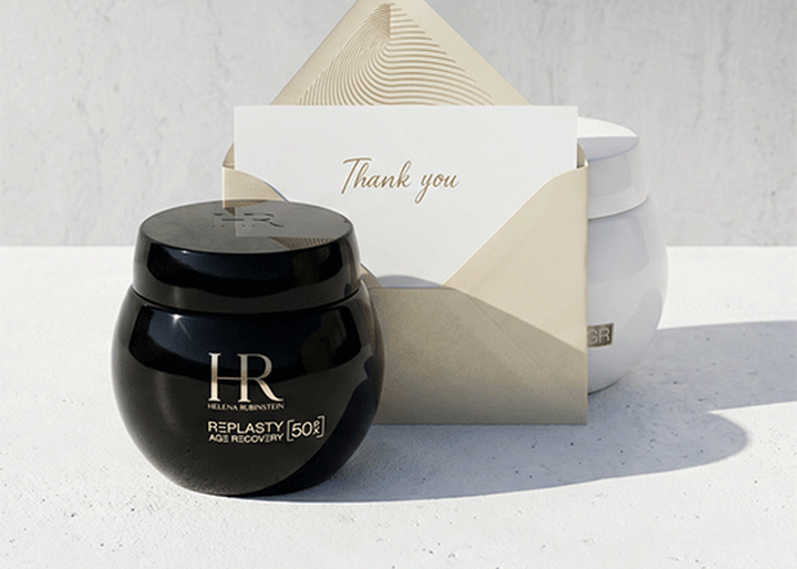 Ein schwarzer Tiegel Helena Rubinstein Replasty Age Recovery Creme und ein Umschlag mit einer „Thank you“-Karte auf einer weißen Oberflache, mit einem weißen Tiegel im Hintergrund.